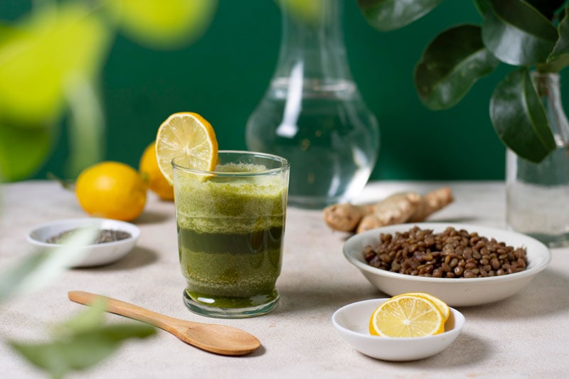 glass of green juice garnished with lemon, next to a bowl of lemon slices and a bowl of seeds