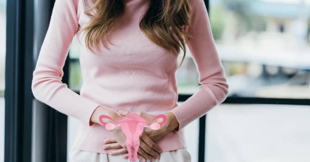 Woman in pink sweater holding a paper uterus over her lower abdomen, symbolizing urinary or pelvic discomfort.